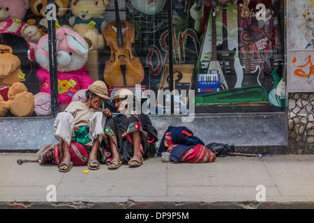 Indigenous man and woman begging while sitting on the sidewalk in front of a store window in Sucre, Bolivia. Stock Photo