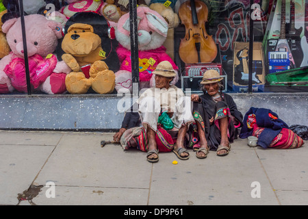 Indigenous man and woman begging while sitting on the sidewalk in front of a store window in Sucre, Bolivia. Stock Photo