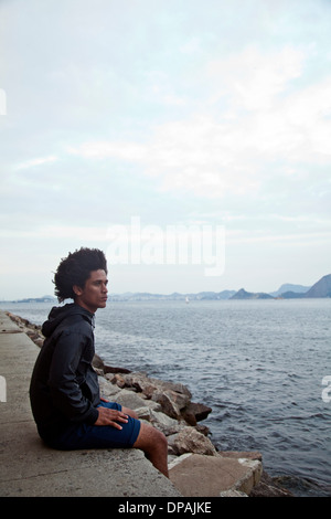 Young man sitting on wall looking at sea, Rio de Janeiro, Brazil Stock Photo