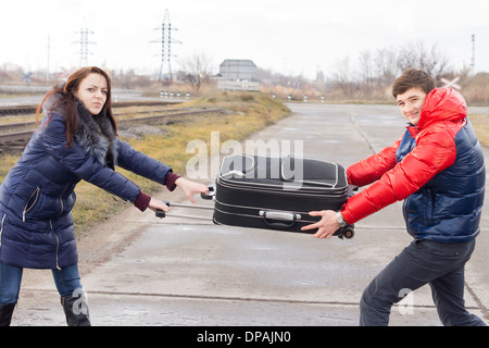 Young man and woman fighting and arguing each pulling on one end of a suitcase as they stand in a rural road alongside a railway line. Stock Photo