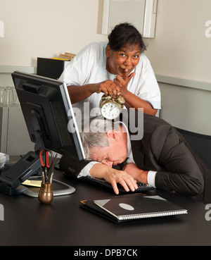 Cleaning woman holding an alarm clock next to a sleeping office worker Stock Photo