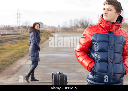 Young couple waiting with their suitcase in the middle of the road on a cold winter day with the young man keeping a sharp lookout for their transport while his girlfriend wanders around behind. Stock Photo