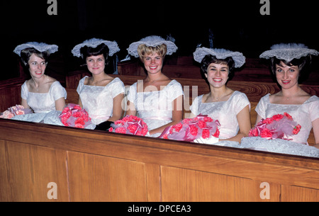 Five smiling bridesmaids in matching white dresses and frilly hats await in a church pew for a 1960s wedding ceremony to begin. Stock Photo