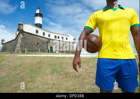 Brazilian football player standing with vintage brown soccer ball at Farol da Barra lighthouse in Salvador Stock Photo