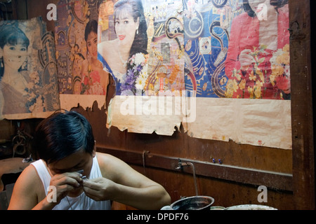 A jeweler is making jewelry in a booth decorated with colorful posters  in the Russian Market in Phnom Penh, Cambodia. Stock Photo