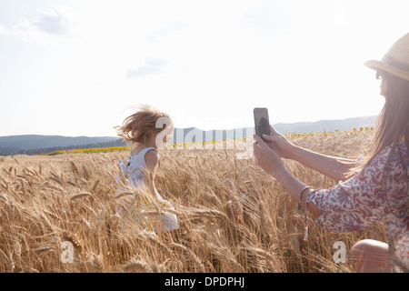 Mother photographing girl running through wheat field Stock Photo