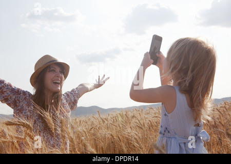 Girl taking photograph of mother in wheat field with arms open Stock Photo