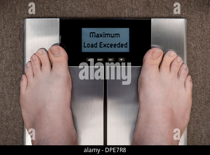 Man's Feet on Bathroom Scales on Carpet photographed from above with Display Showing MAXIMUM LOAD EXCEEDED Stock Photo