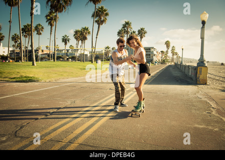 Young woman on skateboard at San Diego beach, boyfriend helping Stock Photo