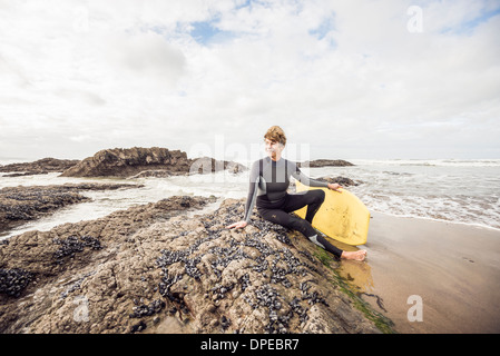 Mature female bodyboarder sitting on rocks, Devon, UK Stock Photo