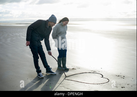 Young couple drawing heart in sand, Brean Sands, Somerset, England Stock Photo