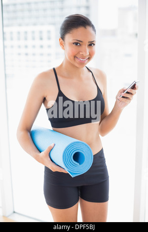 Cheerful dark haired model in sportswear carrying an exercise mat and a mobile phone Stock Photo