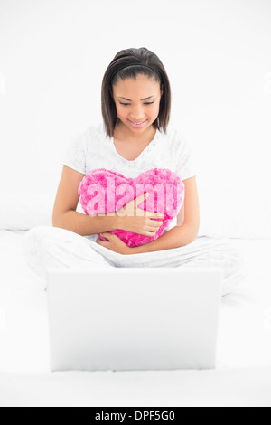 Concentrated young dark haired model cuddling a pillow and looking at a laptop Stock Photo