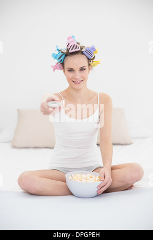 Concentrated natural brown haired woman in hair curlers watching tv while eating popcorn Stock Photo