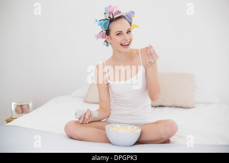 Happy natural brown haired woman in hair curlers watching tv while eating popcorn Stock Photo