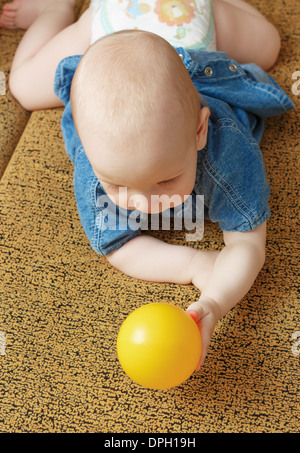 The baby holds a yellow ball lying on a yellow sofa. Stock Photo