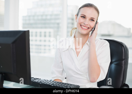 Smiling businesswoman sitting at desk talking on smartphone Stock Photo