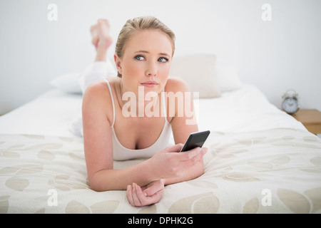 Natural pensive blonde lying on bed and using smartphone Stock Photo