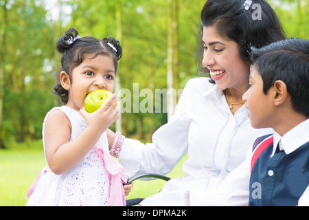 Happy Indian family. Asian girl eating an green apple at outdoor with mother and sibling. Stock Photo