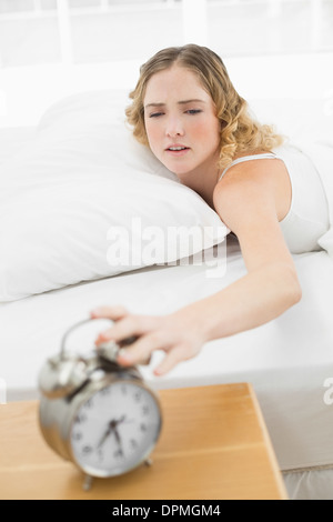 Pretty upset blonde lying in bed turning off alarm clock Stock Photo