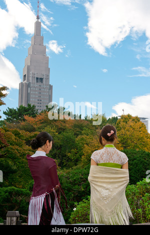 Two young women in traditional Vietnamese attire facing Shinjuku skyscraper behind a wall of trees in a Central Tokyo park. Stock Photo