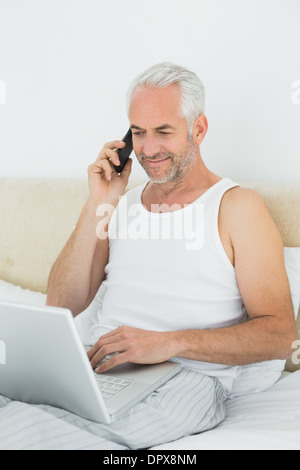 Mature man using cellphone and laptop in bed Stock Photo