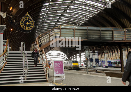 Interior of trainshed with iron & glass roof, stationary trains & woman walking by platform clock - York Railway Station, North Yorkshire, England, UK Stock Photo