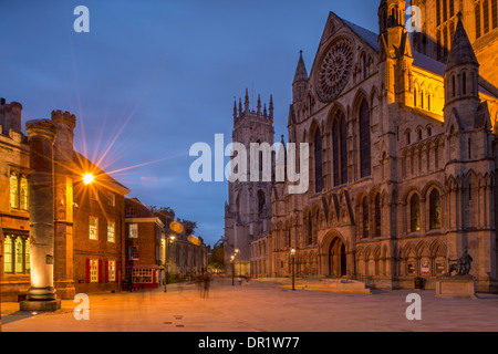 Evening in scenic piazza - south entrance to magnificent illuminated York Minster & blurred figures of people - York, North Yorkshire, England, UK. Stock Photo