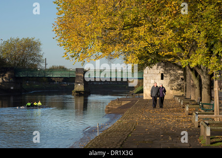 Rowers in boat & couple walking - quiet, scenic, sunlit, tree-lined riverside footpath in autumn - River Ouse, Dame Judi Dench Walk, York, England, UK Stock Photo