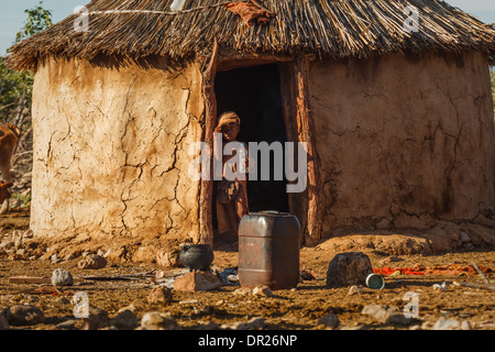 Himba child poses in middle of doorway to mud house with thatched roof in Damaraland, Namibia, Africa Stock Photo