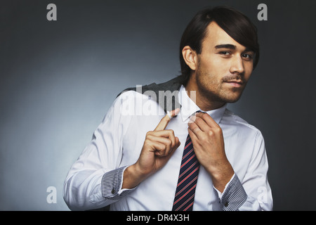 Asian businessman posing against grey background. Young male model holding his jacket and adjusting his necktie. Stock Photo