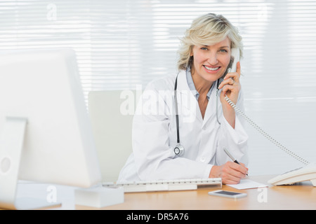 Smiling doctor with computer using phone at medical office Stock Photo