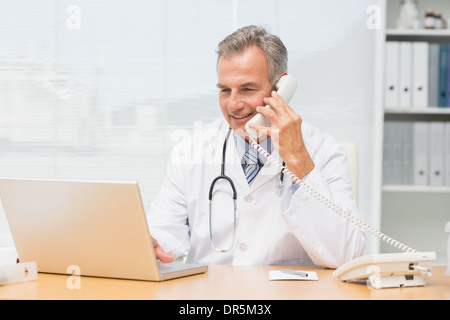 Doctor using laptop and talking on phone at desk Stock Photo
