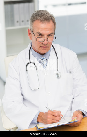 Serious doctor sitting at his desk writing on clipboard Stock Photo