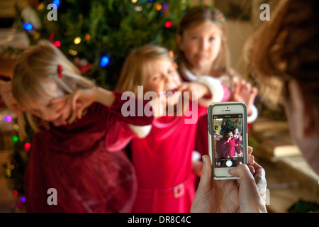 Wearing red holiday clothes, three sisters happily pose for their mother's cell phone camera in front of the family Christmas tree in Laguna Niguel, CA. Stock Photo