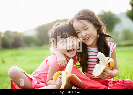 two girls sitting on grass eating bananas Stock Photo