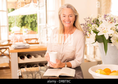 Senior woman drinking coffee and reading book in kitchen Stock Photo