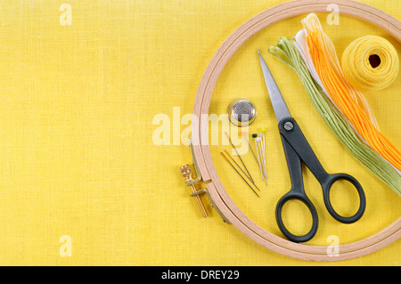 Embroidery and cross stitch accessories on yellow linen fabric. Embroidery hoop, scissors, thread, needles, thimble. Copy space. Stock Photo