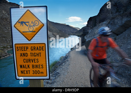 Mountain biker and warning sign on Roxburgh Gorge Cycle and Walking Track, Central Otago, South Island, New Zealand Stock Photo