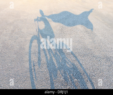 Superhero child wearing a cape riding a bicycle as a cast shadow on a road pretending to be a powerful hero as a metaphor and symbol of youth and childhood imagination and empowering kids self esteem. Stock Photo