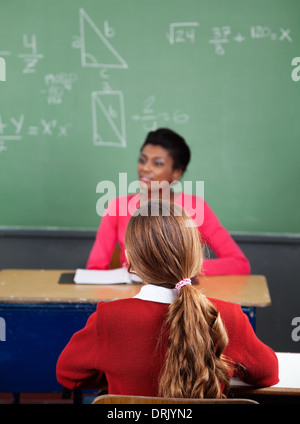 Schoolgirl Sitting At Desk With Teacher In Background Stock Photo