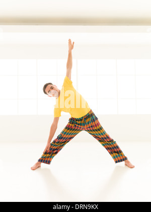 Colorful dressed male repeating Trikonasana yoga exercises in a white room with window background Stock Photo