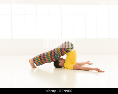 Colorful dressed male repeating Halasana yoga exercises in a white room with window background Stock Photo