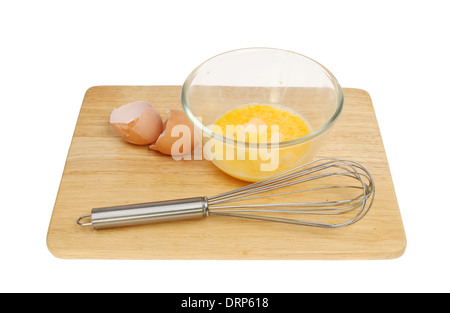 Egg in a glass mixing bowl with a whisk and egg shells on a wooden board isolated against white Stock Photo