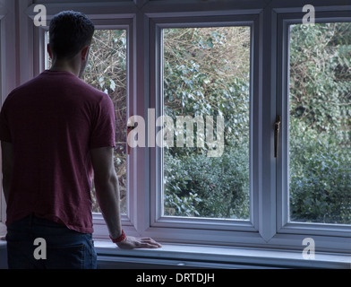 Young man indoors looking out through a window, over shoulder view. Stock Photo