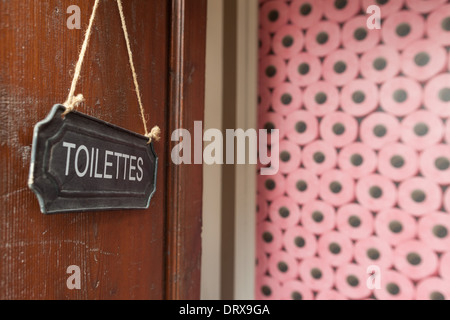 Toilet sign hanging on a wooden doors with stack of pink toilet paper rolls in background. Stock Photo