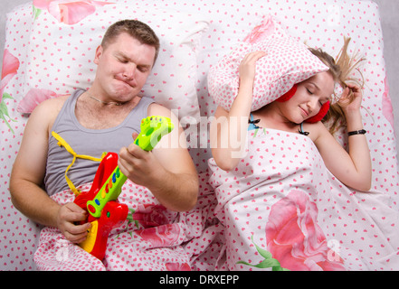 Young couple lying in bed. Husband plays guitar and shouts of his voice. Wife covered her pillow Stock Photo