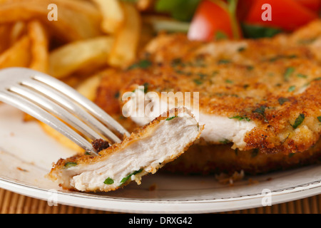 A piece of homemade breaded chicken schnitzel or escalope on a fork with french fries and a tomato and green salad behind Stock Photo