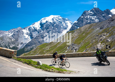 Cyclists ride roadbikes behind motorcycle uphill on The Stelvio Pass, Passo dello Stelvio, Stilfser Joch, in the Alps, Italy Stock Photo