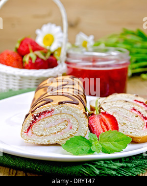 Biscuit roulade with cream and jam, jar of jam, napkin, chamomile, strawberries, mint on the background of wooden boards Stock Photo